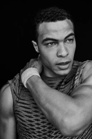 Black and white portrait of a man wearing a patterned sleeveless top and a wristband, looking off to the side.