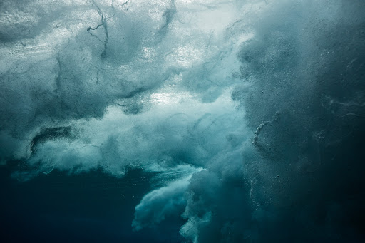 Underwater view of an ocean wave crashing, featuring swirling water and turbulent textures in shades of blue and white.