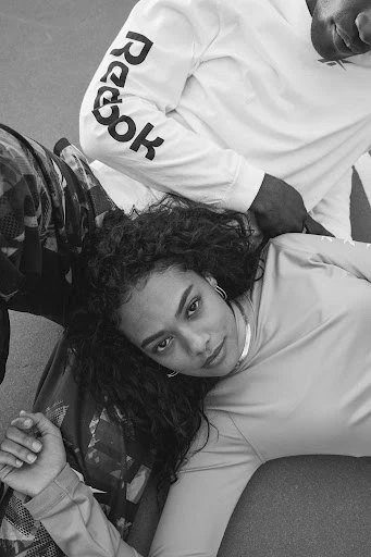 Overhead shot of a young woman with curly hair lying down, wearing a long-sleeved shirt, with a man in a Reebok shirt next to her.