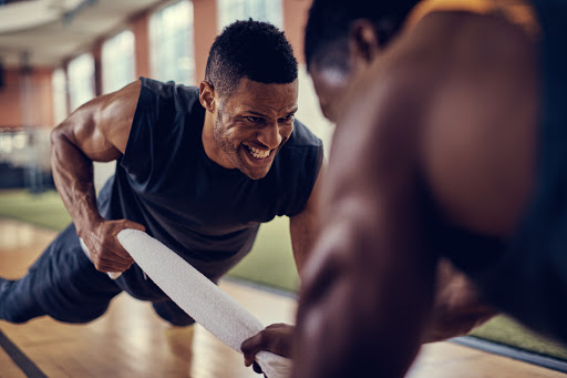 Two muscular men work out together, one in a push-up position with a towel while the other provides resistance, in a fitness center.