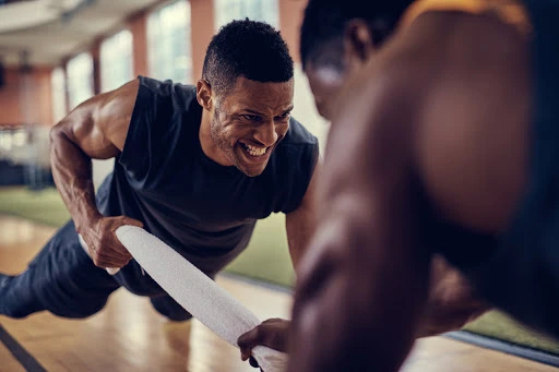 Two muscular men work out together, one in a push-up position with a towel while the other provides resistance, in a fitness center.