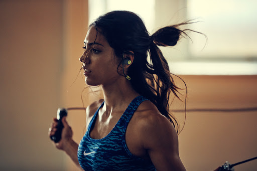 Athletic woman in workout clothes jumps rope in a gym, wearing earbuds and a Nike top, showcasing fitness and active lifestyle.