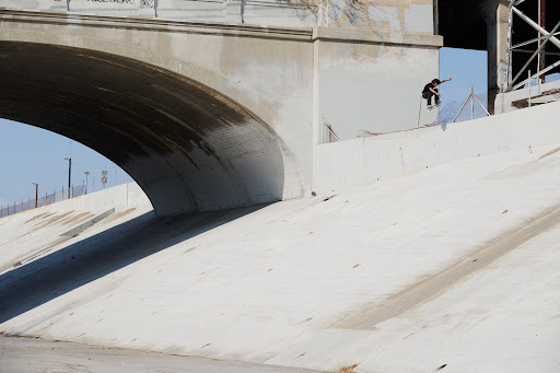 A skateboarder performs a trick on a concrete ledge under a bridge, with a clear sky and urban backdrop.