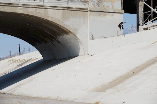 A skateboarder performs a trick on a concrete ledge under a bridge, with a clear sky and urban backdrop.