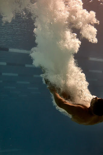Underwater view of adult male diving into a pool, creating bubbles and motion as he enters the clear blue water.