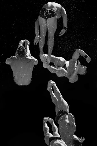 Monochromatic underwater view of five adult male divers in mid-dive with dark background and water droplets visible, emphasizing athletic skill.