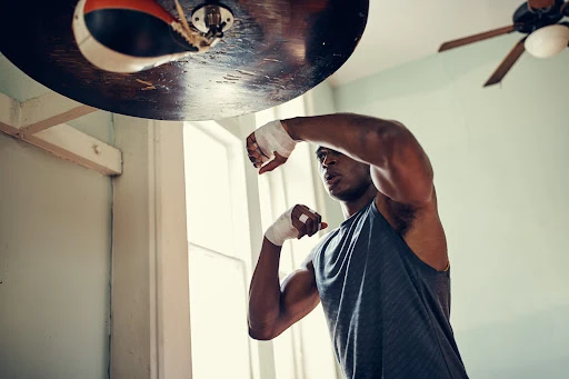 A muscular man with boxing wraps on his hands practices punching a speed bag in an indoor workout space during the daytime.
