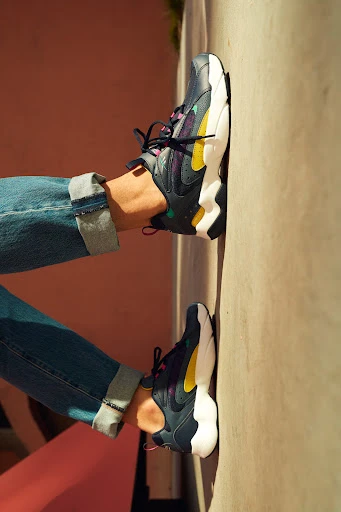 Close-up of a person wearing blue jeans and blue, yellow, and white athletic shoes leaning against a beige wall.