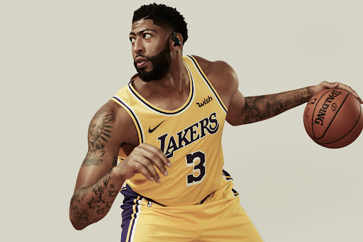 Basketball player in Lakers jersey dribbling a Spalding ball on a neutral background; he's wearing wireless headphones and has multiple tattoos.