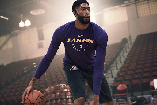 A professional basketball player, Anthony Davis, dribbles a basketball on an indoor court, wearing a Los Angeles Lakers long-sleeve shirt.