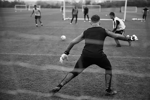 Black and white shot of soccer goalie in action, with other players and goals in the background, set on a grassy field.