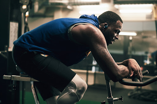 Sweaty adult male on stationary bike at gym, wearing blue hooded athletic top and compression pants, looking down with focused expression.