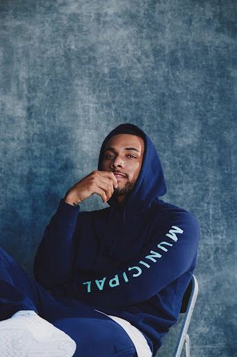 A man in a hooded sweatshirt, with MUNICIPAL printed vertically on the sleeve, poses in a studio against a textured background.