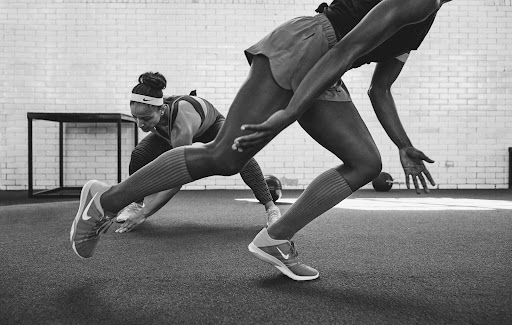 Two athletic women in workout gear perform floor exercises with a medicine ball in a gym with white brick walls in a monochrome image.