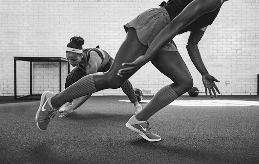 Two athletic women in workout gear perform floor exercises with a medicine ball in a gym with white brick walls in a monochrome image.