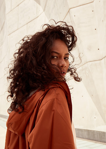 Woman with long dark curly hair wearing an orange hooded jacket looking back over her shoulder outdoors against a geometric concrete wall