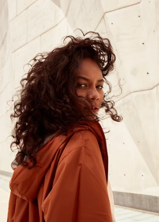 Woman with long dark curly hair wearing an orange hooded jacket looking back over her shoulder outdoors against a geometric concrete wall