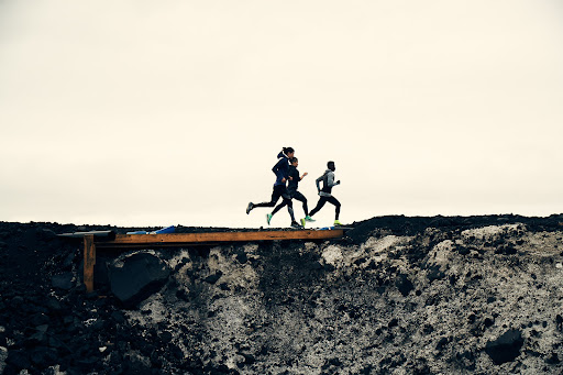 A group of four athletes run in unison along a narrow wooden bridge atop a craggy, dark-gray ridge against a flat, light sky.