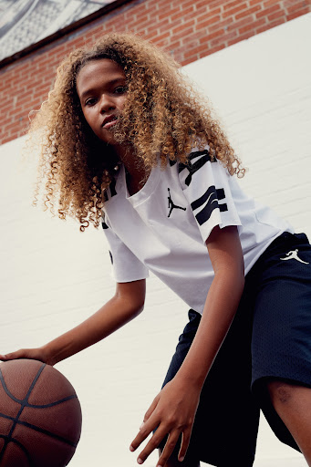 Young person dribbling a basketball outdoors in front of brick wall wearing a white t-shirt and black shorts with logos.