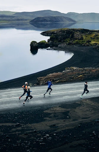 Group of runners on a road next to a lake and volcanic landscape. Overcast day, athletic wear, running shoes, and natural scenery.