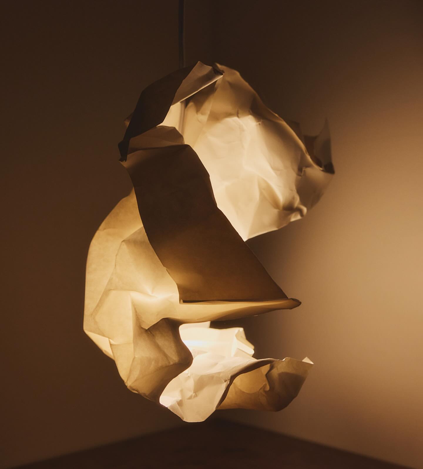 Abstract hanging lamp sculpture made from light brown crumpled paper lit from within, casting a warm glow in a darkened room.