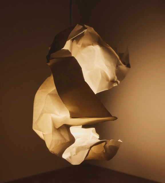 Abstract hanging lamp sculpture made from light brown crumpled paper lit from within, casting a warm glow in a darkened room.