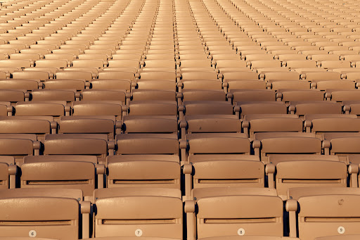 Rows of empty tan-colored stadium seats stretch into the distance, creating a symmetrical and repetitive pattern under even lighting.