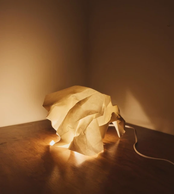 Indoor shot of a crumpled paper lamp casting a warm glow on a wooden surface against a plain wall in a corner, creating a cozy ambiance.
