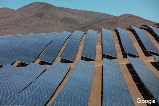 Aerial view of a large solar panel farm in a desert landscape, creating clean, renewable energy with mountains in the background under a clear blue sky.