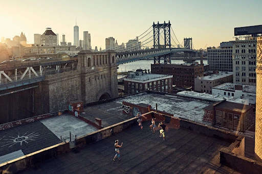 Four women perform choreographed dance on a rooftop overlooking the Manhattan Bridge and New York City skyline at dusk or dawn.