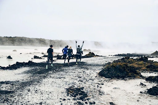 Four athletic adults running on a volcanic rock landscape near geothermal waters on an overcast day; activewear and outdoor fitness.