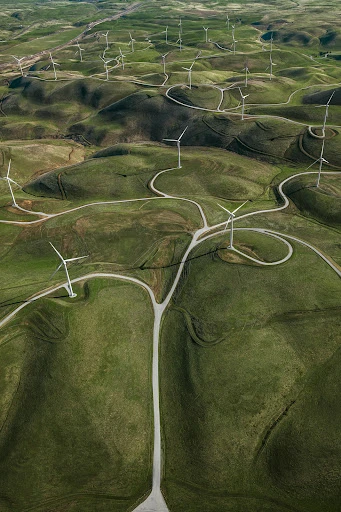 Aerial view of a wind farm featuring numerous wind turbines on rolling green hills, connected by a network of dirt roads.
