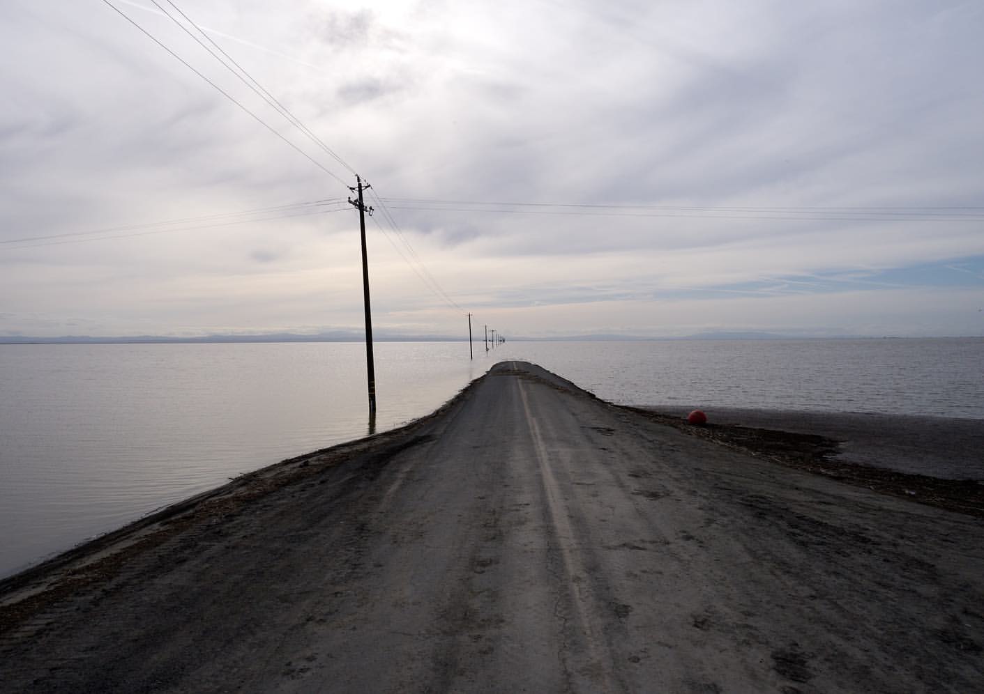 An eye-level landscape photograph showcases a road submerged in water, stretching into the horizon under a cloudy sky, with power poles.