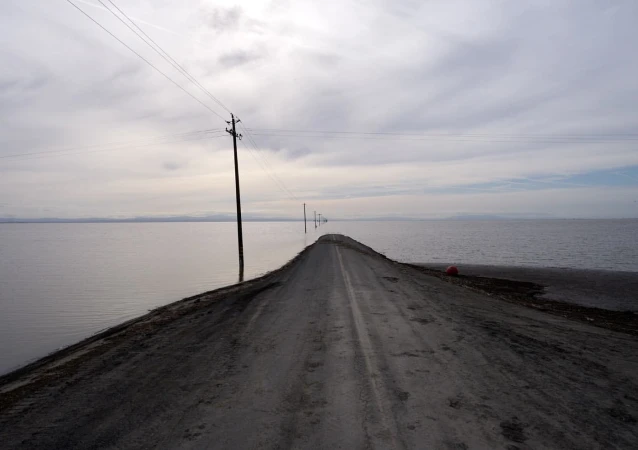 An eye-level landscape photograph showcases a road submerged in water, stretching into the horizon under a cloudy sky, with power poles.