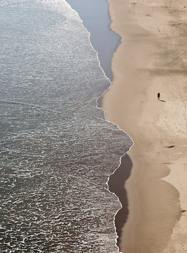 Aerial view of a lone person walking along a wide sandy beach beside gentle ocean waves breaking on the shore.