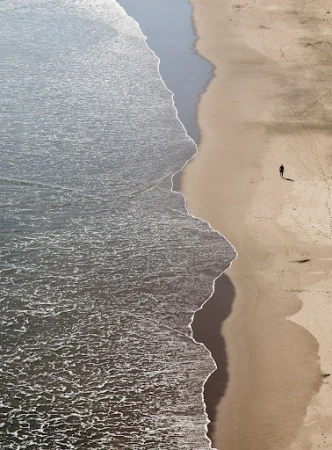 Aerial view of a lone person walking along a wide sandy beach beside gentle ocean waves breaking on the shore.