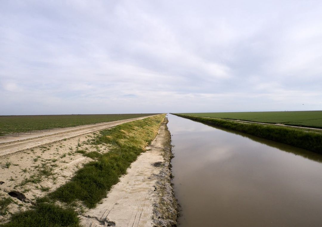 Scenic view of a canal cutting through a rural landscape, reflecting the cloudy sky, with cultivated fields on either side.