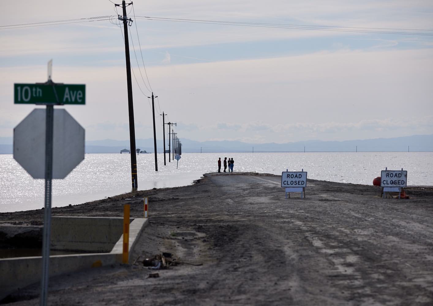 Group of people standing on a road flooded with water, with utility poles, road closed signs, and distant mountains in the background.