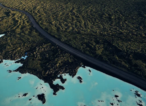 Aerial view captures a stark black road winding through a desolate green landscape and next to light-blue water with black rocks.