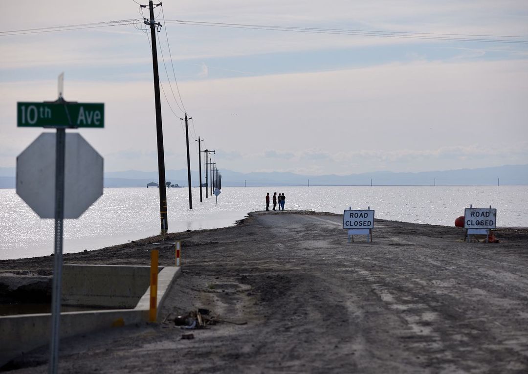 Flooded road with utility poles disappearing into standing water under a cloudy sky, with a group of people on the horizon.