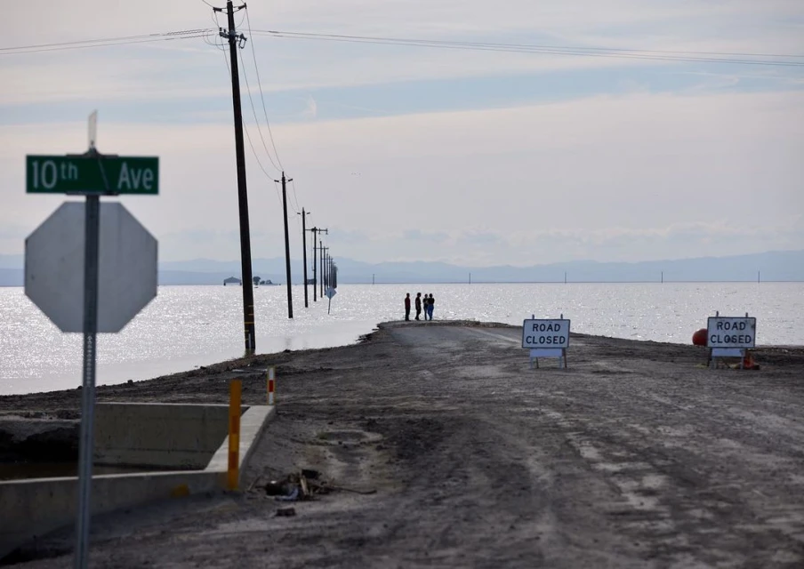 Flooded road with utility poles disappearing into standing water under a cloudy sky, with a group of people on the horizon.