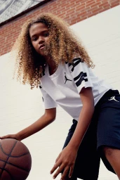 Young person with curly blonde hair dribbling a basketball outdoors against a brick and white wall