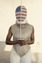 Fencer wearing a mask painted with the American flag, holding a foil upright against a plain white background.