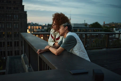 Two young adults leaning on a rooftop railing at dusk, overlooking city buildings.
