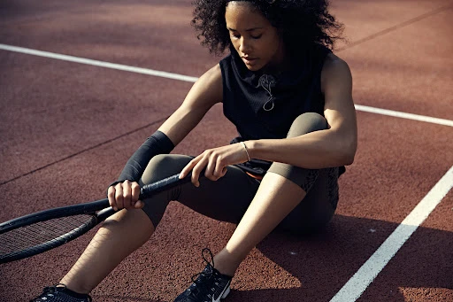 Woman sitting on a tennis court holding a racket while wearing athletic wear.
