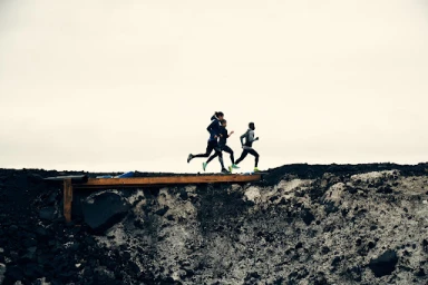 Three runners in athletic wear sprinting across a dark terrain under an overcast sky, photographed by Sam Kweskin.