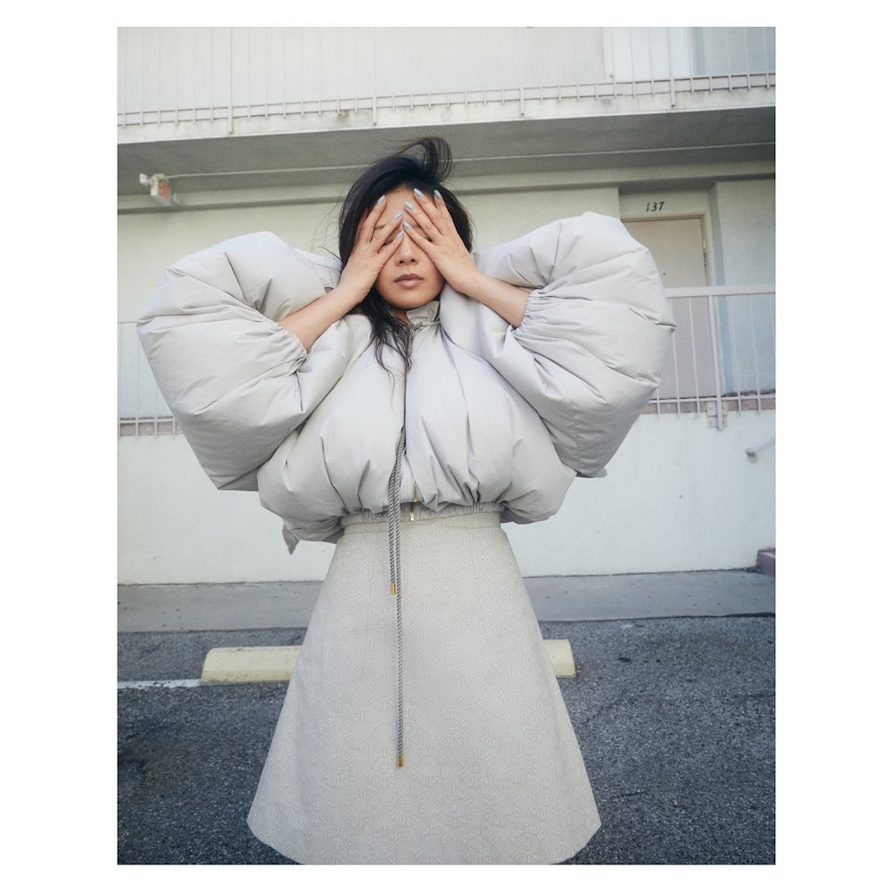 Young woman with dark hair poses outdoors in a padded jacket and skirt, covering her eyes with her hands. Building in background.