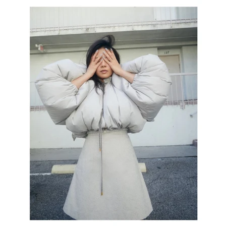Young woman with dark hair poses outdoors in a padded jacket and skirt, covering her eyes with her hands. Building in background.