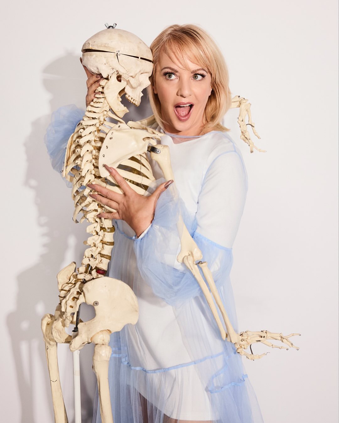 A fair-skinned woman with blonde hair strikes a playful pose with an anatomical skeleton against a white background in a studio setting.