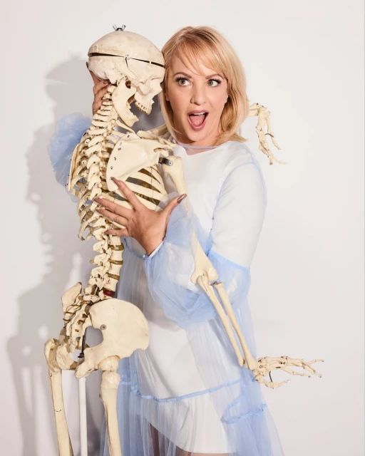 A fair-skinned woman with blonde hair strikes a playful pose with an anatomical skeleton against a white background in a studio setting.
